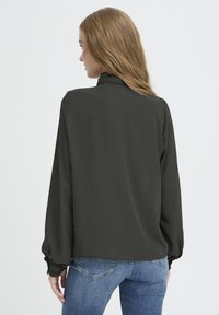 Dunkelgrüne Bluse mit langen, gerafften Ärmeln und einem hohen, rüschierten Kragen. Getragen über blauen Jeans, mit einer glatten, weichen Stoffstruktur.