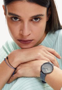 Zilveren horloge met een ronde wijzerplaat en een gestreept blauwe band, gedragen om een pols. Het horloge heeft een grijze wijzerplaat met dunne uurmarkeringen.