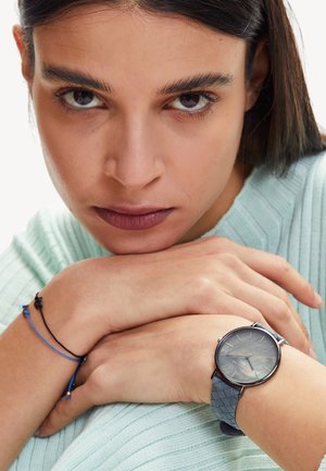 Montre argentée avec un cadran rond et un bracelet bleu à motifs, portée au poignet. La montre possède un cadran gris avec de fins index horaires.