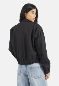 Czarna kurtka bomberka o skróconym kroju, z elastycznym pasem i długimi rękawami, wykonana z gładkiego materiału. Połączona z jasnoniebieskimi dżinsami.