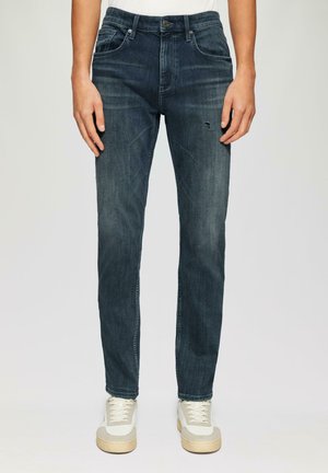 Jean en denim bleu foncé à coupe slim, avec cinq poches, une légère décoloration et une petite déchirure usée sur la cuisse avant gauche.