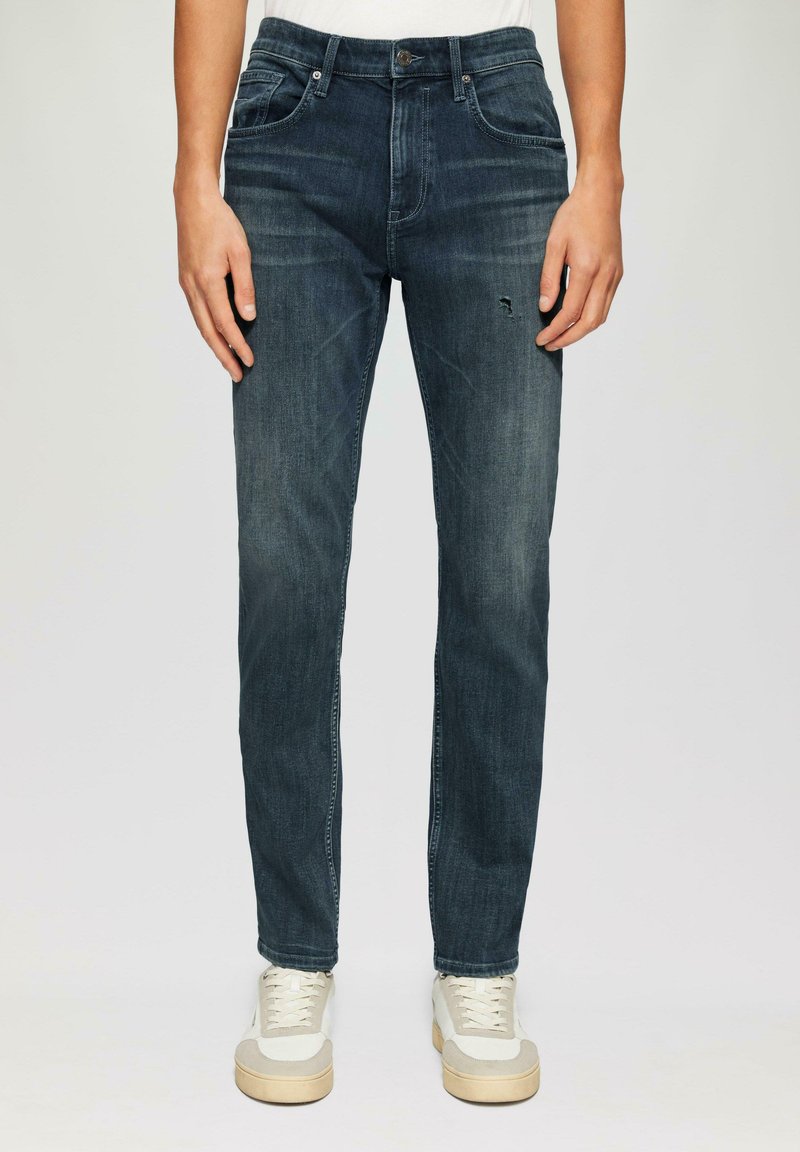Jeans in denim blu scuro con una vestibilità slim, dotati di cinque tasche, leggero sbiadimento e un piccolo strappo consumato sulla parte anteriore della coscia sinistra.