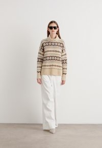 Pull en tricot beige avec un motif, col haut et ourlet côtelé, assorti à un pantalon large blanc et des chaussures beiges.