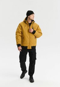 Mężczyzna w musztardowym żółtym długim kapturze, czarnych spodniach cargo, czarnych butach i czapce beanie, stoi i patrzy w lewo.