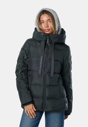Jeune femme portant une doudoune noire avec un grand col, une capuche en tricot grise et un jean bleu, debout devant un fond uni.