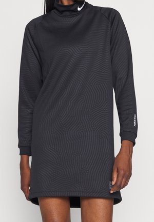 Frau trägt ein schwarzes langärmliges Nike-Kleid mit dezenten Strukturmustern, einem hohen Kragen und dem Schriftzug "NIKE FC." auf dem Ärmel.