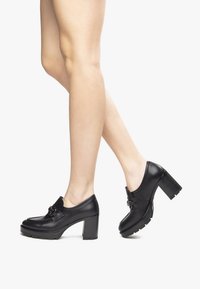 Scarpe nere in pelle con tacco, punta rotonda, una prominente fibbia decorativa e un tacco spesso texturizzato, che mostra una superficie liscia.