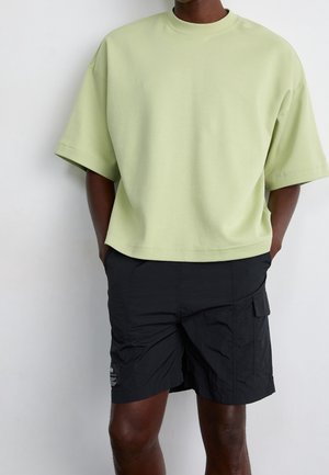 Mand iført lys grøn oversized kortærmet skjorte og sorte cargoshorts står med hænderne i lommerne.