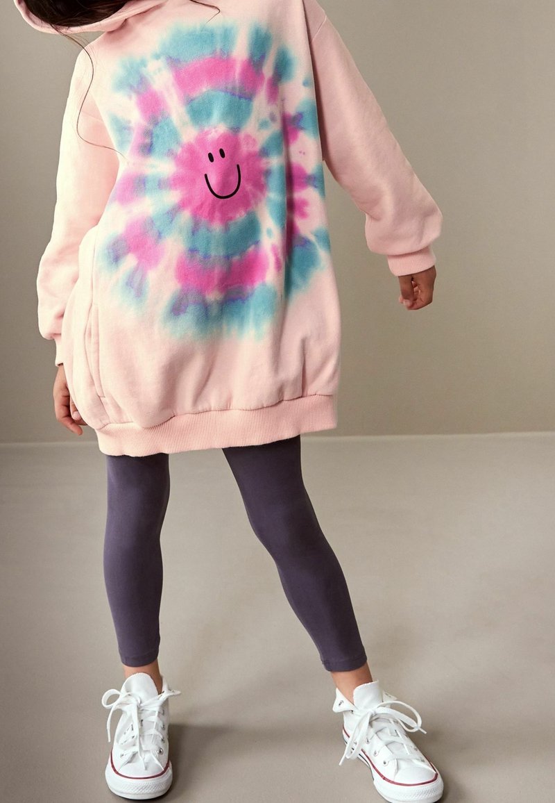 Enfant portant un sweat à capuche rose surdimensionné avec un visage souriant tie-dye bleu et rose, des leggings gris foncé et des baskets blanches, debout sur un sol beige.