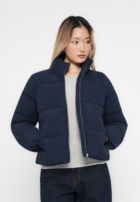 Ung kvinne iført en marineblå polstret jakke over en lys grå skjorte og mørke jeans, ser mot siden med hendene i jakkelommene.