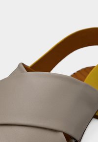 Δερμάτινο σανδάλι ανοιχτού γκρι χρώματος με επικαλυπτόμενους ιμάντες. Κίτρινα και καφέ στοιχεία στη σόλα. Ομαλή υφή, με περίγραμμα σχήματος.