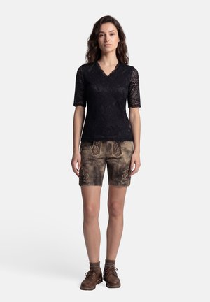 Frau steht und trägt ein schwarzes Spitzen-Oberteil mit kurzen Ärmeln, bestickte braune Lederhosen, braune Socken und braune Schnürschuhe vor weißem Hintergrund.