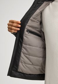 Schwarze Lederjacke, geöffnet, um ein graues, gestepptes Innenfutter mit einer kleinen Reißverschlusstasche zu zeigen. Ärmel- und Kragenverzierungen sind sichtbar.