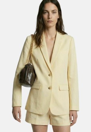 Mujer con blazer amarillo pálido y pantalones cortos a juego, llevando un pequeño bolso marrón oscuro con cadena dorada en el hombro.