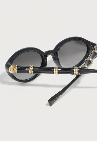 Μαύρα οβάλ γυαλιά ηλίου με σκούρους φακούς, βραχίονες σε στυλ μπαμπού διακοσμημένοι με χρυσές λεπτομέρειες και το "GUCCI" τυπωμένο στην άκρη των βραχιόνων.