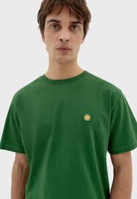Camiseta de algodón verde con cuello redondo, mangas cortas y un pequeño logo de sol bordado en el lado izquierdo del pecho. Textura suave.