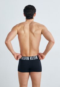 Sorte boxer truser med en hvit Calvin Klein-logo på linningen, vist fra baksiden på en bart overkropp med en glatt hudtekstur.
