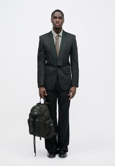 Μαύρο κοστούμι με γιακά σερβιέτας σε πράσινο χρώμα, πουκάμισο με σχέδια και γραβάτα. Κρατάει ένα μαύρο σακίδιο με πολλές τσέπες και ιμάντες. Μαύρα παπούτσια.