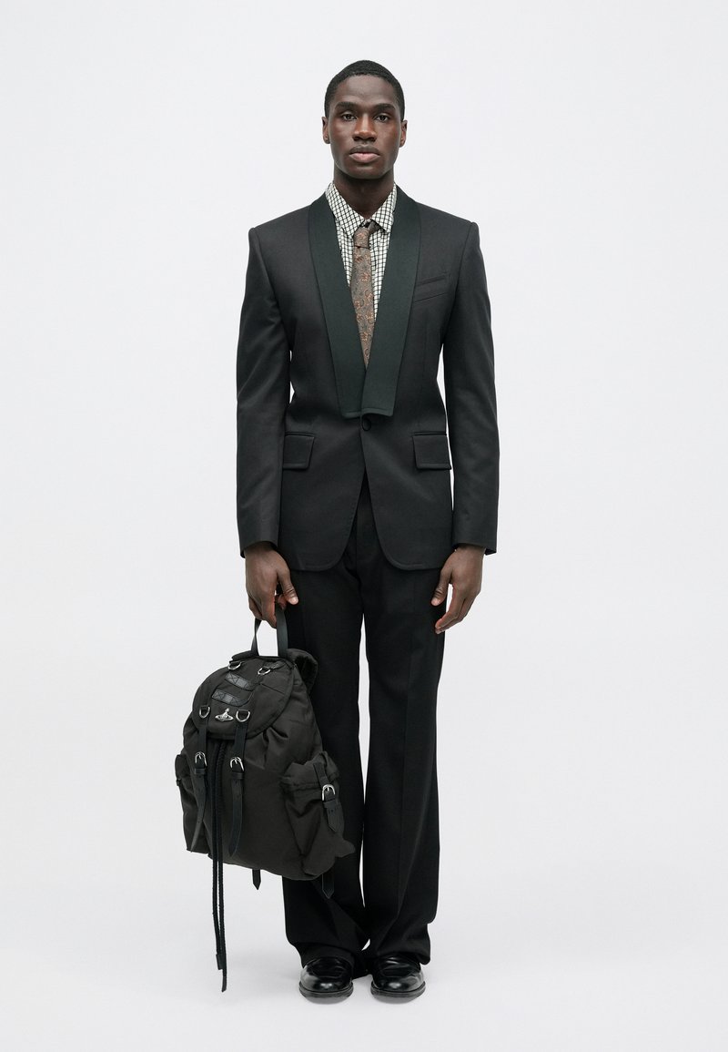 Sort jakkesæt med grøn sjalskrave, mønstret skjorte og slips. Holder en sort rygsæk med flere lommer og stropper. Sorte sko.