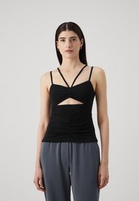 Top negro sin mangas con un diseño recortado, frente fruncido y tirantes delgados. Tela suave con una silueta ajustada. Combinado con pantalones de pierna ancha grises.