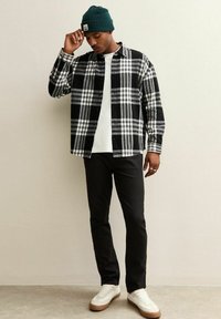 Camicia di flanella a scacchi in bianco e nero, indossata sopra una maglietta bianca, abbinata a pantaloni neri e sneakers bianche. Gli accessori includono un berretto verde.