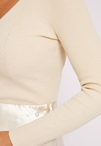 Beige geribbelde lange mouwen top met een V-hals en textuurdetails, gedragen met een zijden crèmekleurige tailleband met een zilveren "G" accent.