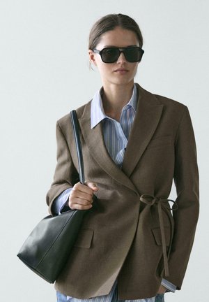 Donna che indossa un blazer marrone sopra una camicia a righe blu, occhiali da sole neri, con una borsa nera appoggiata su una spalla, su sfondo chiaro.