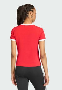 Czerwona koszulka z krótkim rękawem z białym wykończeniem, o dopasowanym kroju i gładkiej fakturze, widziana od tyłu. Czarne spodnie z widocznymi paskami.