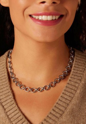 Zilveren metalen ketting met onderling verbonden cirkelvormige en ovale schakels. Glad oppervlak met een gepolijste afwerking, gedragen om de nek boven een beige trui.