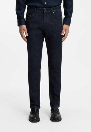 Hombre vistiendo vaqueros ajustados azul oscuro, zapatos de cuero negros y una camisa oscura, de pie contra un fondo blanco liso.