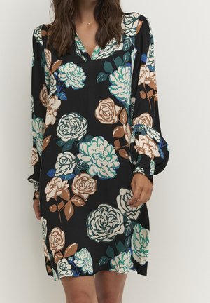 Femme portant une robe noire à manches longues avec de grands motifs floraux blancs, verts et beige et des poignets smockés, debout devant un fond uni.