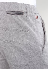 Pantalon en tissu tissé gris avec une coupe droite, comportant une poche arrière et une étiquette de marque cousue à la taille. Accent rouge près de la poche.