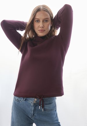 Burgunderfarbener Pullover mit höherem Kragen, langen Ärmeln und einem verstellbaren Kordelzugsaum. Aus einem strukturierten Stoff gefertigt, getragen mit blauen Jeans.