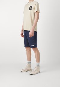 Camiseta de algodón beige con bolsillo de logo negro, pantalones cortos azul marino y zapatillas altas beige con acentos grises. Textura lisa y diseño sencillo.