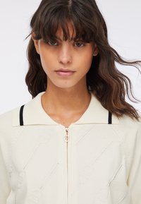 Giovane donna con capelli scuri e mossi che indossa un maglione crema con zip, colletto e un motivo fosse in rilievo sottile, rivolta in avanti su uno sfondo bianco.