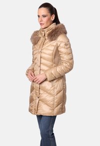 Mujer con un abrigo de invierno acolchado beige y un gran cuello de piel, de pie con las manos entrelazadas, vistiendo jeans azules frente a un fondo liso.