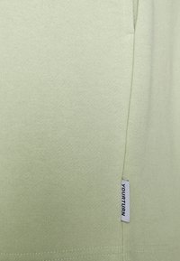 Světle zelené tričko vyrobené z texturované látky s jemnými detaily šití, na boku s malou bílou nálepkou s nápisem "TVŮJ OTOČ".