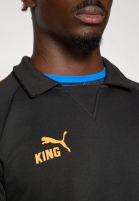 Sort polo shirt med krave, med orange "KING" tekst og Puma logo. Underneath ses en blå skjorte med et diskret mønster.