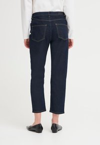 Persona que lleva puesto un pantalón de mezclilla recto de color azul oscuro con bolsillos traseros, una blusa blanca y zapatos negros de tacón bajo, de pie contra un fondo claro y liso.