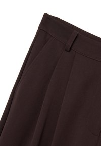 Pantalones marrones ajustados con un frente plano, que presentan dos pliegues laterales, una textura suave y una cinturilla con pasadores.