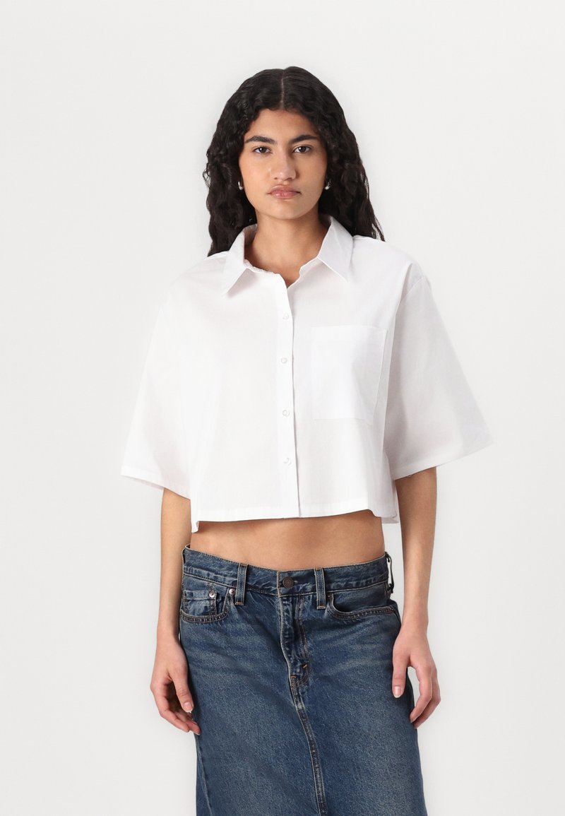 Camisa branca cropped com botões, de mangas curtas e um bolso no peito, combinada com uma saia de ganga azul. Textura suave, design limpo.
