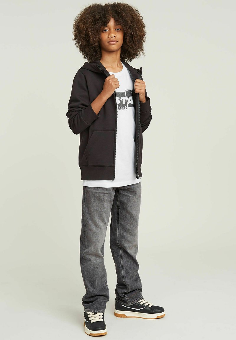 Sort zip-up hættetrøje med sidelommer, iført over en hvid grafisk t-shirt. Grå jeans og sorte sneakers med en hvid sål fuldender outfittet.