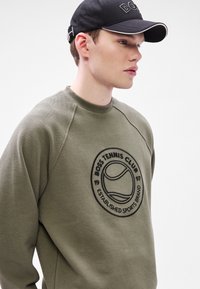 Zelená mikina s okrúhlym čiernym logom s tenisovým loptičkovým vzorom. Materiál má textúru, mikina má okrúhly výstrih a raglánové rukávy.