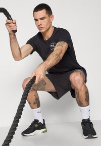 Sorte atletiske shorts, sort t-shirt med logo og sorte sneakers med grønne accenter. Engagerer sig med et tykt, tekstureret kampreb.