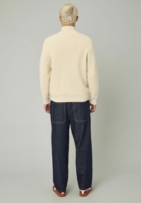 Pull en maille côtelée crème avec col haut, associé à un jean en denim bleu large avec poches arrière visibles et coutures contrastées.