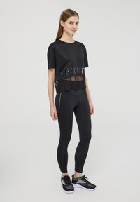 Top atletico nero con orlo trasparente e testo stampato, abbinato a leggings neri con strisce bianche ai lati e scarpe da ginnastica nere con rete.