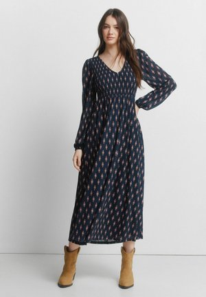 Donna in piedi che indossa un vestito lungo a maniche lunghe blu navy con un piccolo motivo e stivali color cuoio alla caviglia, su uno sfondo chiaro e semplice.