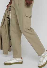 Person, der beiges Cargo-Hosengesäß und weiße Sneakers mit braunen Akzenten trägt, hält eine passende beige Jacke und geht vor einem einfarbigen Hintergrund.