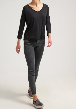 Personne marchant portant un haut noir à manches longues avec col en V, un jean skinny foncé et des chaussures grises sans lacets avec un motif texturé.