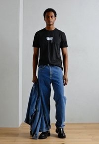 Camiseta negra de manga corta con un logotipo plateado, combinada con jeans de mezclilla azules, botas negras y sosteniendo una chaqueta de mezclilla. Fondo de suelo de madera clara.